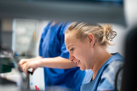 Junge Frau in blauer Arbeitskleidung lächelt konzentriert bei der Arbeit im Labor, während eine Kollegin im Hintergrund arbeitet.