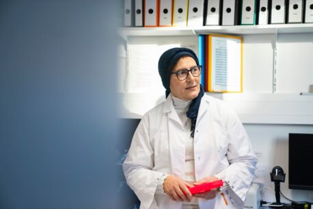 Frau in weißem Laborkittel mit Kopftuch hält einen roten Probenhalter, stehend vor Aktenregalen in einem Laborraum.