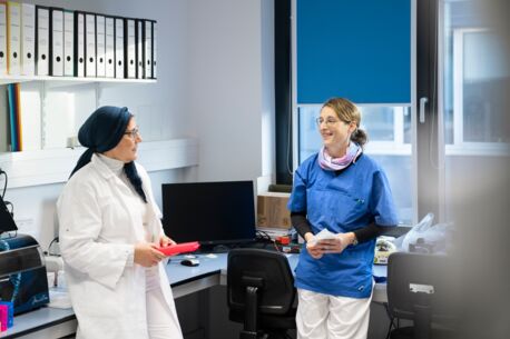 Zwei Frauen, eine im weißen Laborkittel und die andere in blauer Arbeitskleidung, stehen im Laborbüro und unterhalten sich, umgeben von Aktenordnern und Laborgeräten.