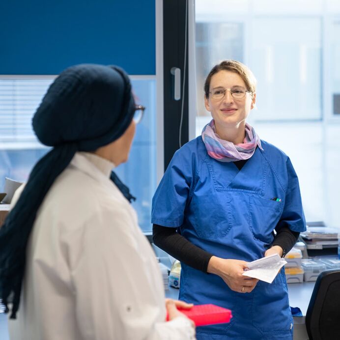 Zwei Frauen im Labor, eine in weißem Laborkittel und die andere in blauer Arbeitskleidung, unterhalten sich, während sie Laborproben und Unterlagen halten.