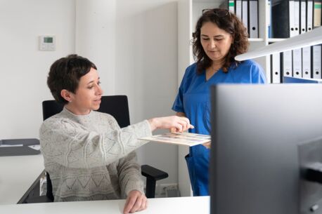 Zwei Frauen im Büro, eine in blauer Arbeitskleidung, die andere in einem Pullover, diskutieren über Dokumente auf einem Tisch.