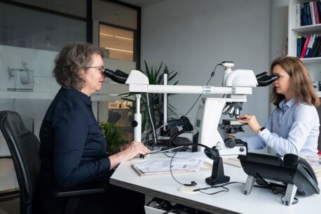 Zwei Frauen in einem modernen Labor sitzen sich gegenüber und arbeiten an einem Mikroskop. Eine Frau trägt ein dunkelblaues Oberteil, die andere ein hellblaues Hemd. Im Hintergrund sind Bücherregale und Pflanzen zu sehen.